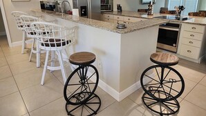 A kitchen with white bar stools, a refrigerator, microwave, and a dishwasher. - Naples Inn & Docks (Naples)