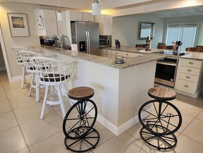 A kitchen with white bar stools, a refrigerator, microwave, and a dishwasher. - Naples Inn & Docks (Naples)