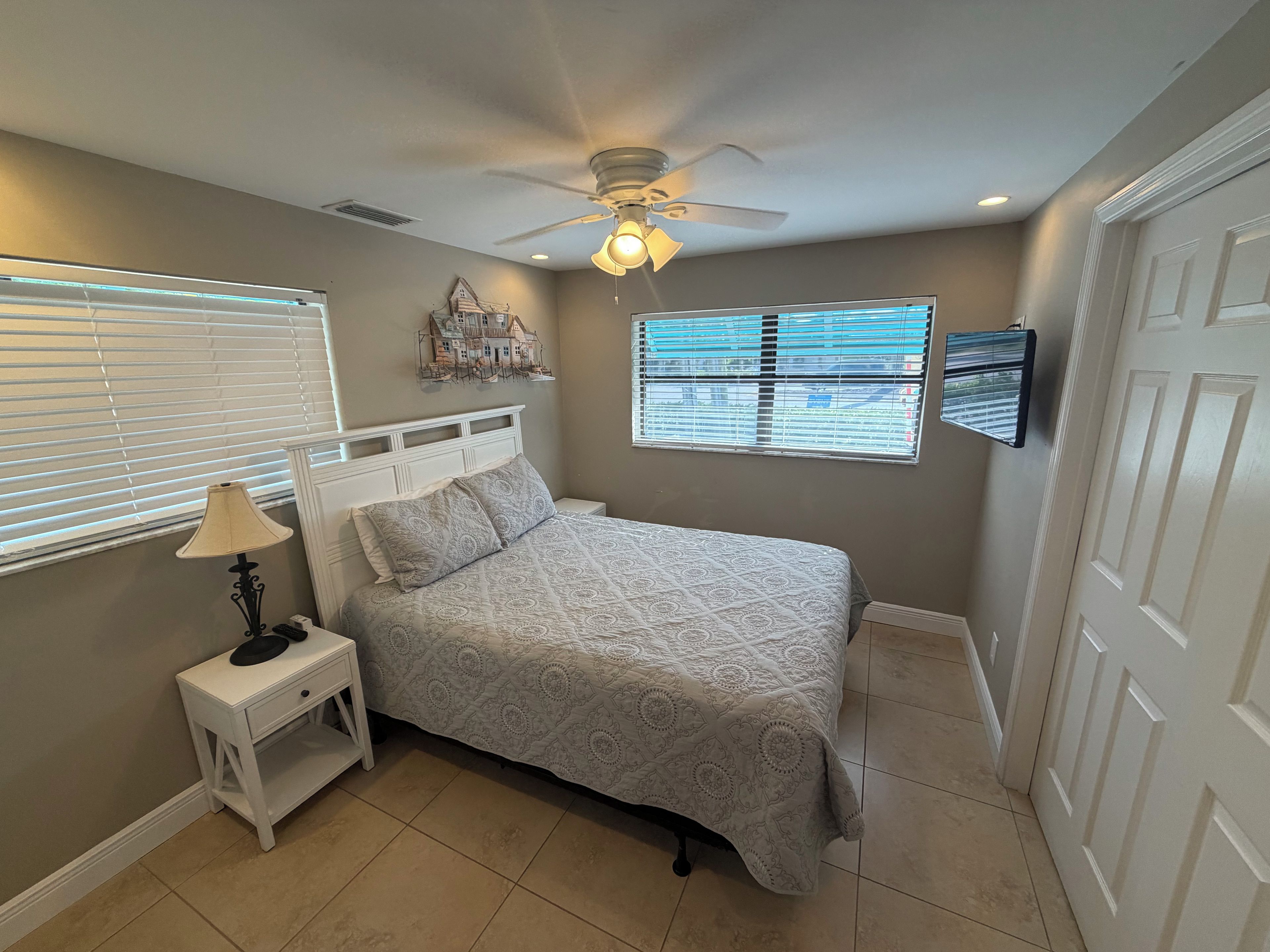 A bedroom with a bed, nightstand, lamp, ceiling fan, window blinds, and a wall-mounted decoration.