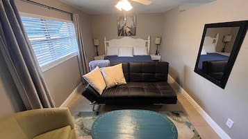 Ein Schlafzimmer mit einem Bett, einem Sofa, einem Sessel, einem runden Tisch, einem Deckenventilator und einem Fenster mit Jalousien.