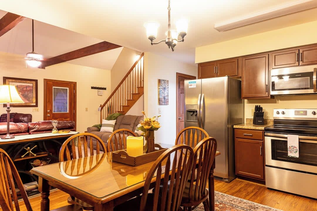 A dining area with a wooden table and chairs, a chandelier, and a kitchen with stainless steel appliances.