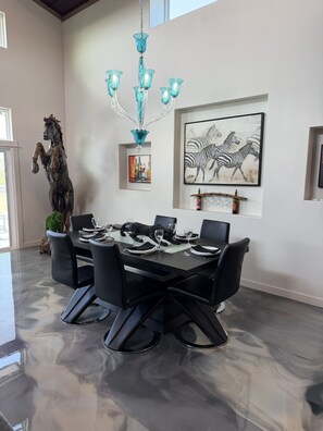 A dining area with a black table set for six, surrounded by black chairs, a blue chandelier, and a zebra painting on the wall. - Wine Down in Blanco — Two King Master Suites & Hill Country Luxury (Blanco)