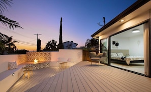 Una terraza moderna al aire libre con zona de estar, un dormitorio con paredes de vidrio y vistas al atardecer.