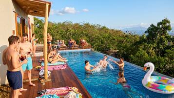 A pool area with people relaxing and swimming, surrounded by greenery and a wooden deck.