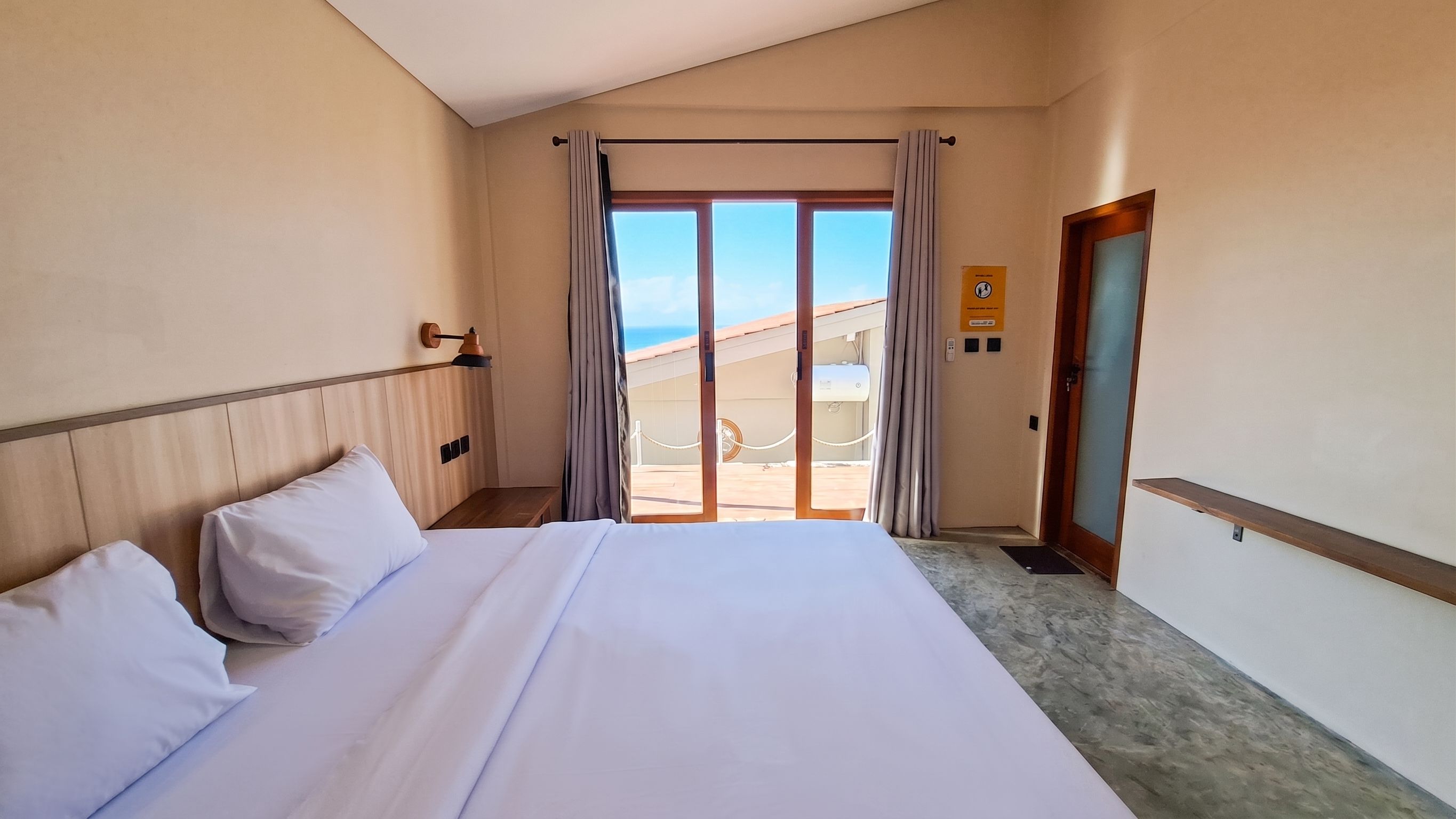 A hotel room with a bed, a nightstand, a lamp, a view of the beach, and a door with a sign.