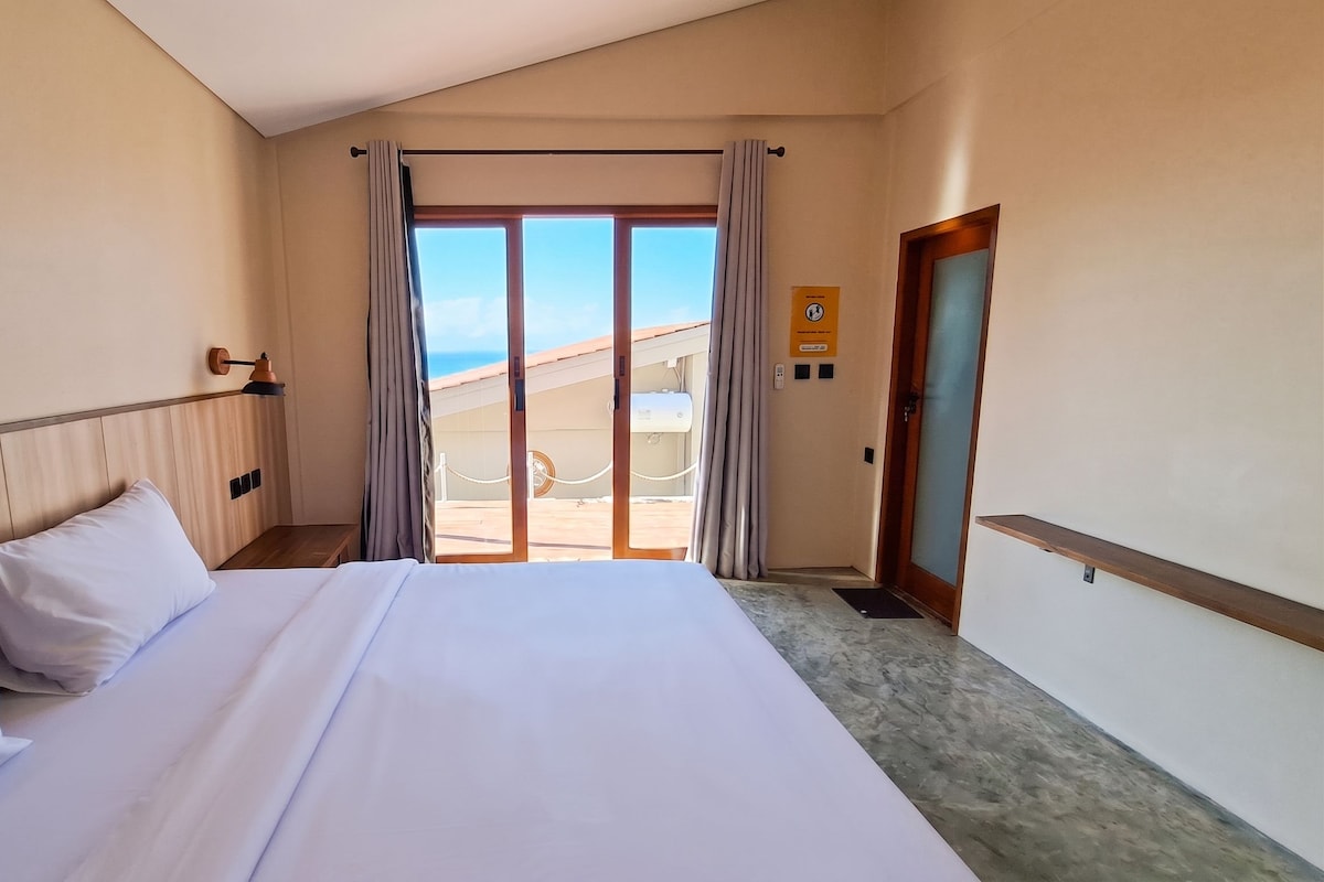 A hotel room with a bed, a nightstand, a lamp, a view of the beach, and a door with a sign.