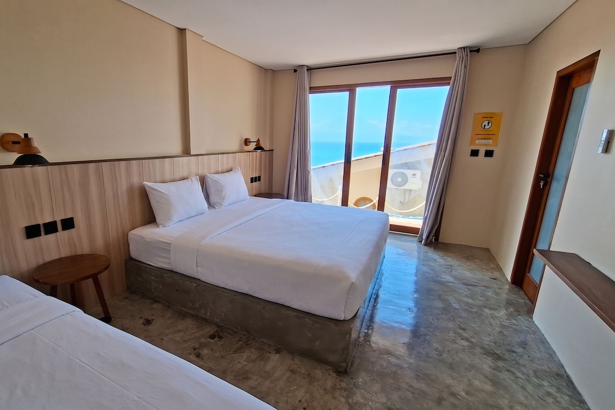 Ein Hotelzimmer mit zwei Betten, einem kleinen Tisch, Meerblick von einem Balkon und einer Tür mit Griff.
