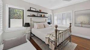 Un dormitorio con cama, mesita de noche, lámpara, ventana con persianas, ventilador de techo y estantería.