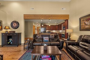 Ein geräumiges Wohnzimmer mit Ledersofas, einem Couchtisch und Holzboden. Im Hintergrund ist eine Küche mit Holzausbau und einem Kühlschrank erkennbar.