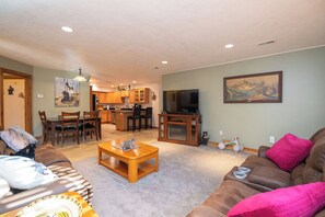A living room with a sofa, coffee table, and a television. There is a dining area with chairs and a table. The kitchen is visible in the background.