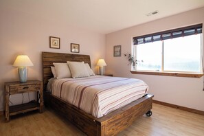 A bedroom with a wooden bed, two bedside tables with lamps, a window with blinds, and framed artwork on the wall.