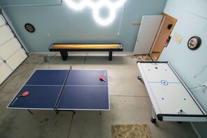 A ping pong table with two paddles, a foosball table, and a dartboard in a room with a concrete floor.