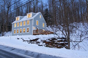 A two-story house with a stone foundation, a wooden deck, and a snow-covered landscape. - Vermont Group Getaway. Hot Tub, Game Room & Reservoir Views. Near Mt Snow (Whitingham)