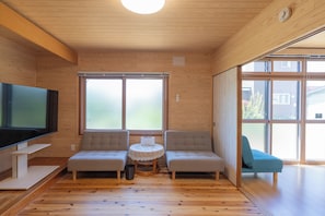 Una sala de estar moderna con un televisor de pantalla plana, dos sillones, una mesita redonda y ventanas amplias.