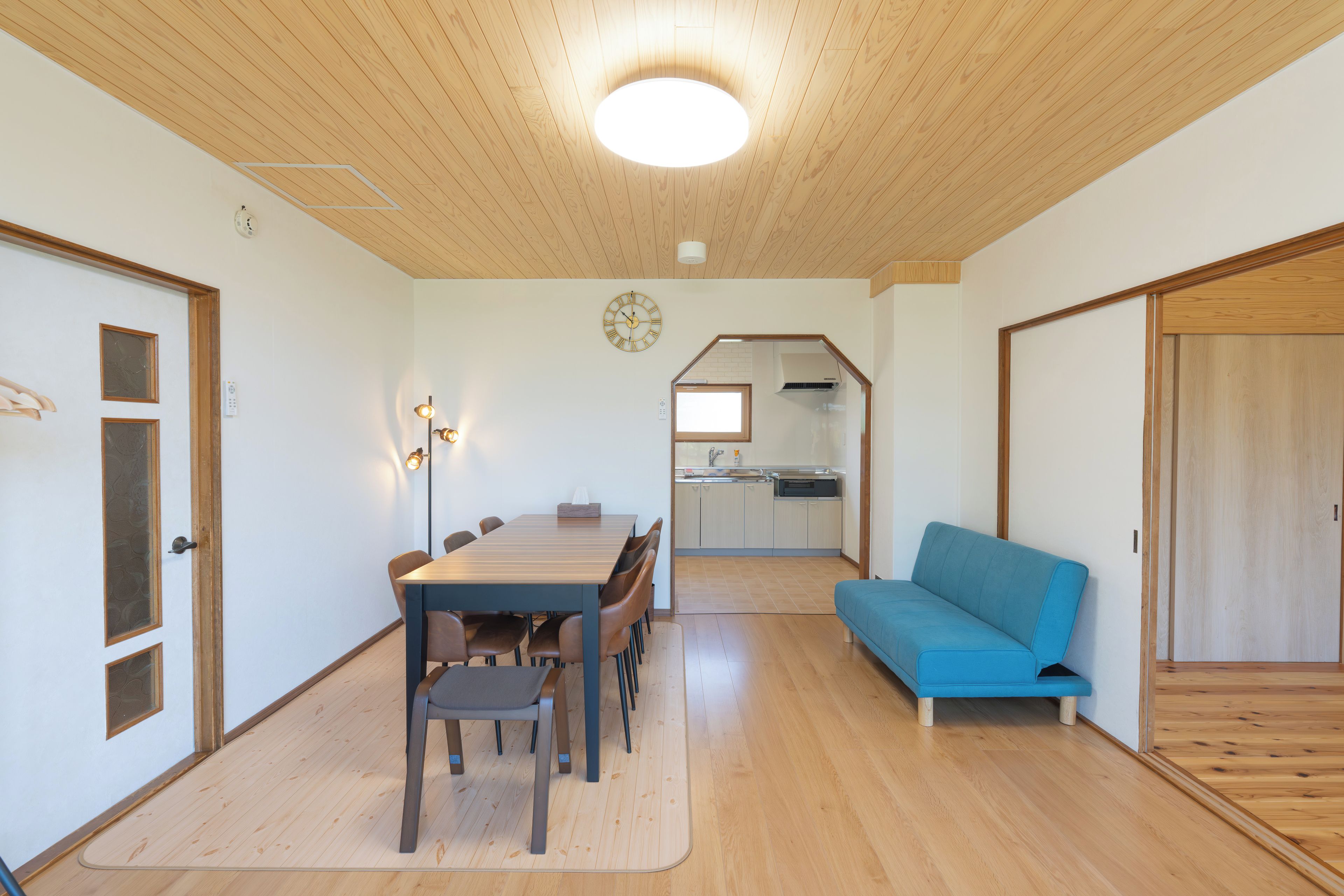 A room with a dining table, chairs, a sofa, and a kitchen area in the background.