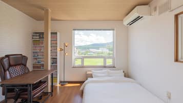 Un dormitorio con cama, escritorio con silla, estantería, ventana con vistas a zonas verdes y edificios, y un equipo de aire acondicionado.