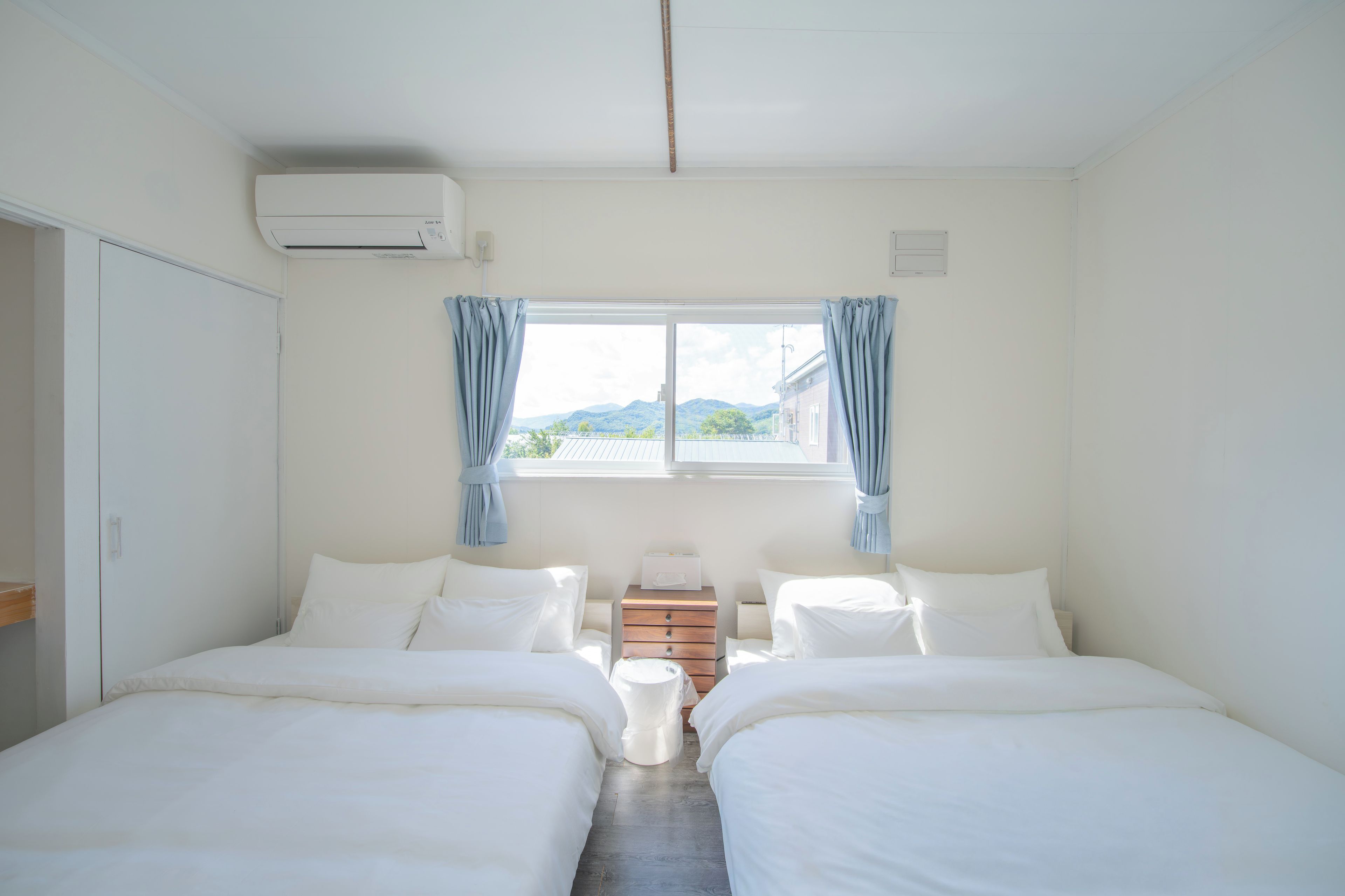 Habitación doble con dos camas, ventana con cortinas, mesita de noche y unidad de aire acondicionado.