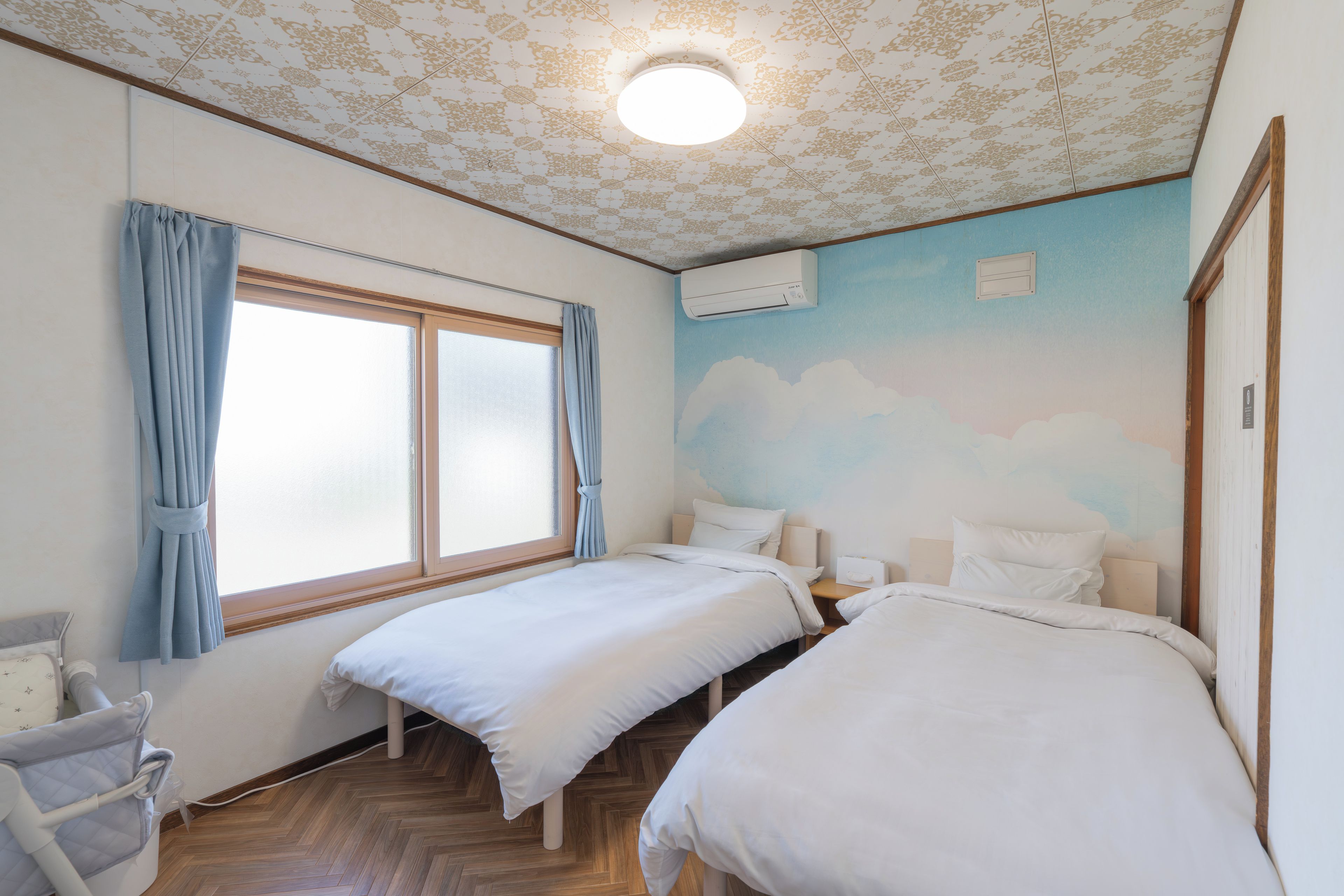 A room with two beds, a window with curtains, a wall-mounted air conditioner, and a ceiling with a floral pattern.