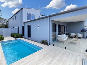 Una casa moderna con piscina, un patio cubierto con mobiliario exterior y un gran ventanal.