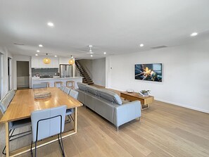 Una cocina moderna con una isla central, un comedor con mesa y sillas de madera, un sofá gris y una escalera que lleva a un nivel superior.