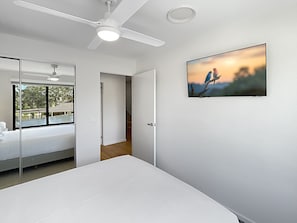 A modern bedroom with a large bed, a ceiling fan, a window with a view, and a wall-mounted TV.