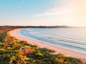 Uma praia com uma linha costeira curva, uma pessoa caminhando na areia e uma vista distante de colinas.