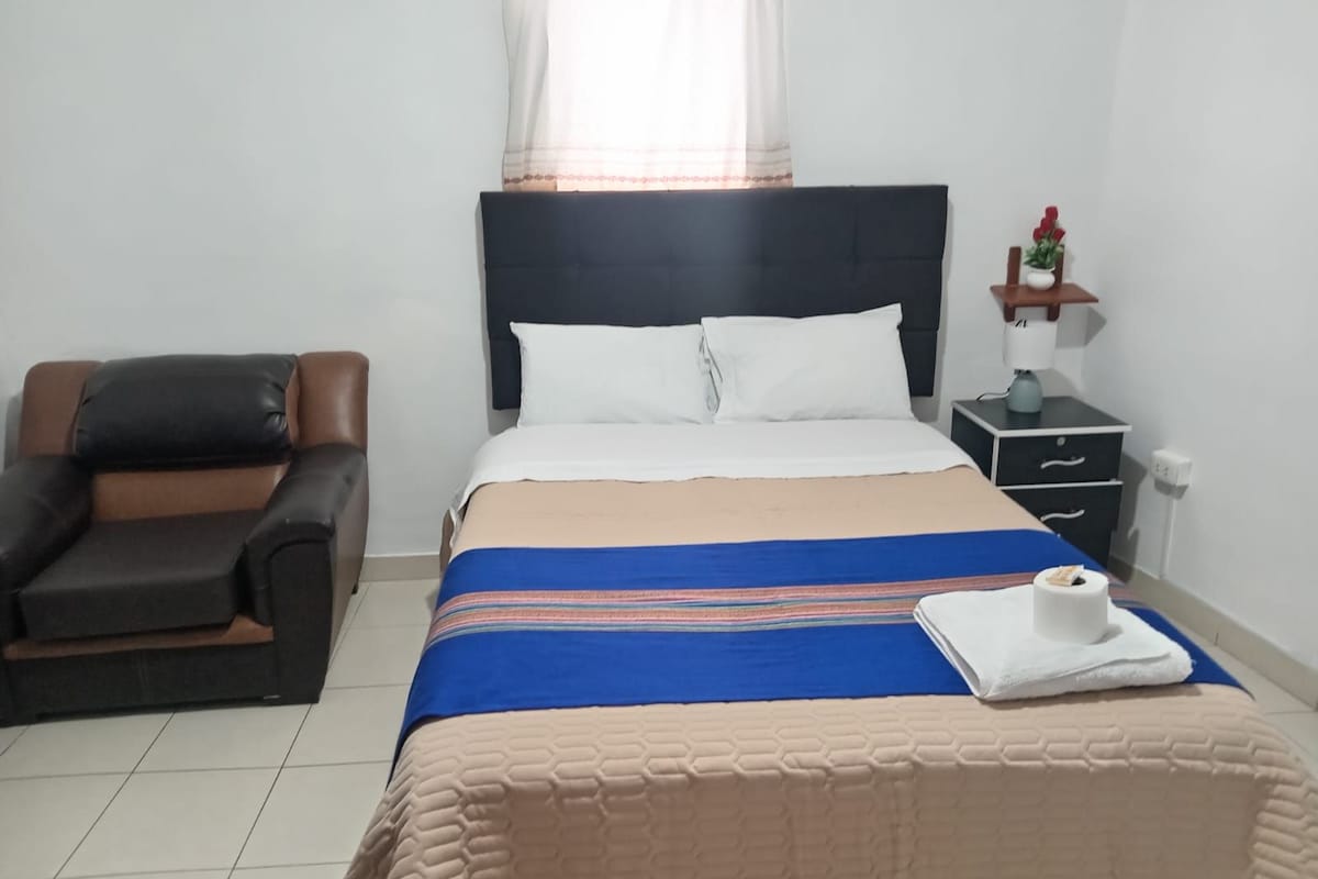 A hotel room with a bed, a chair, a nightstand, and a window with curtains.