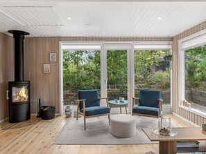 A cozy room with a wood-burning stove, two blue armchairs, a round table, and large windows offering a view of greenery. - "Dirtje" - 850m from the sea by Interhome (Ebeltoft)