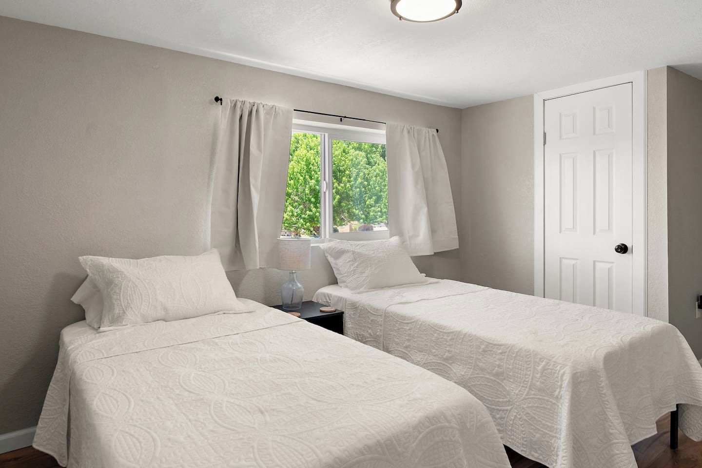 Ein Zimmer mit zwei Betten, einem Fenster mit Vorhängen, einer Tür und einem Nachttisch mit Lampe.
