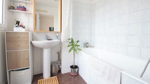 Une salle de bain moderne avec un lavabo blanc, un miroir et une baignoire. On y trouve des étagères avec des articles de toilette et une plante en pot.