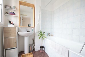 Un baño moderno con lavamanos, espejo y bañera blancos. Hay repisas con artículos de aseo personal y una planta en maceta.