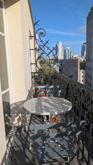 Un balcón con una mesa y sillas de metal, plantas en macetas y vistas de edificios de la ciudad.