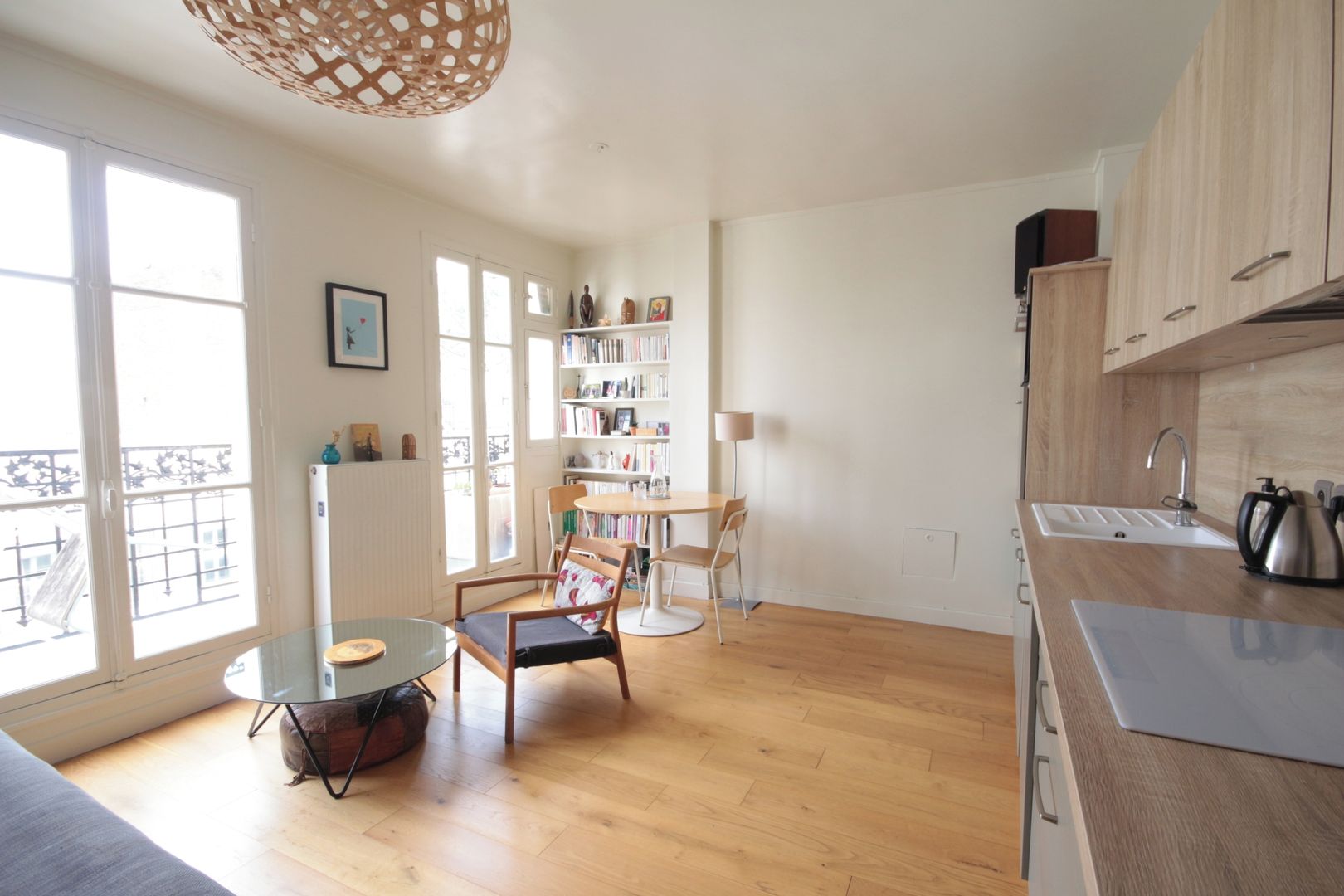 A modern kitchen with wooden cabinets, a dining area with chairs, a bookshelf, and a large window providing natural light.