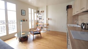 Une cuisine moderne avec des meubles en bois, un coin repas avec des chaises, une bibliothèque et une grande fenêtre qui laisse entrer la lumière naturelle.