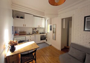 A compact kitchen with white cabinets, a wooden dining table, and a gray sofa in a bright room with a brick wall. - Renovated 1-Bed – Montmartre Village (18th) (Paris)