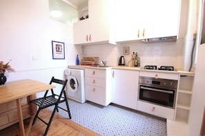 A compact kitchen with a washing machine, oven, and a dining area with a chair and table. - Renovated 1-Bed – Montmartre Village (18th) (Paris)