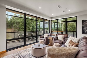 A spacious living room with large windows, leather furniture, and a view of a forested area. - Top-floor corner condo l Hot Tub / Summer Pool / Gym (Whitefish)