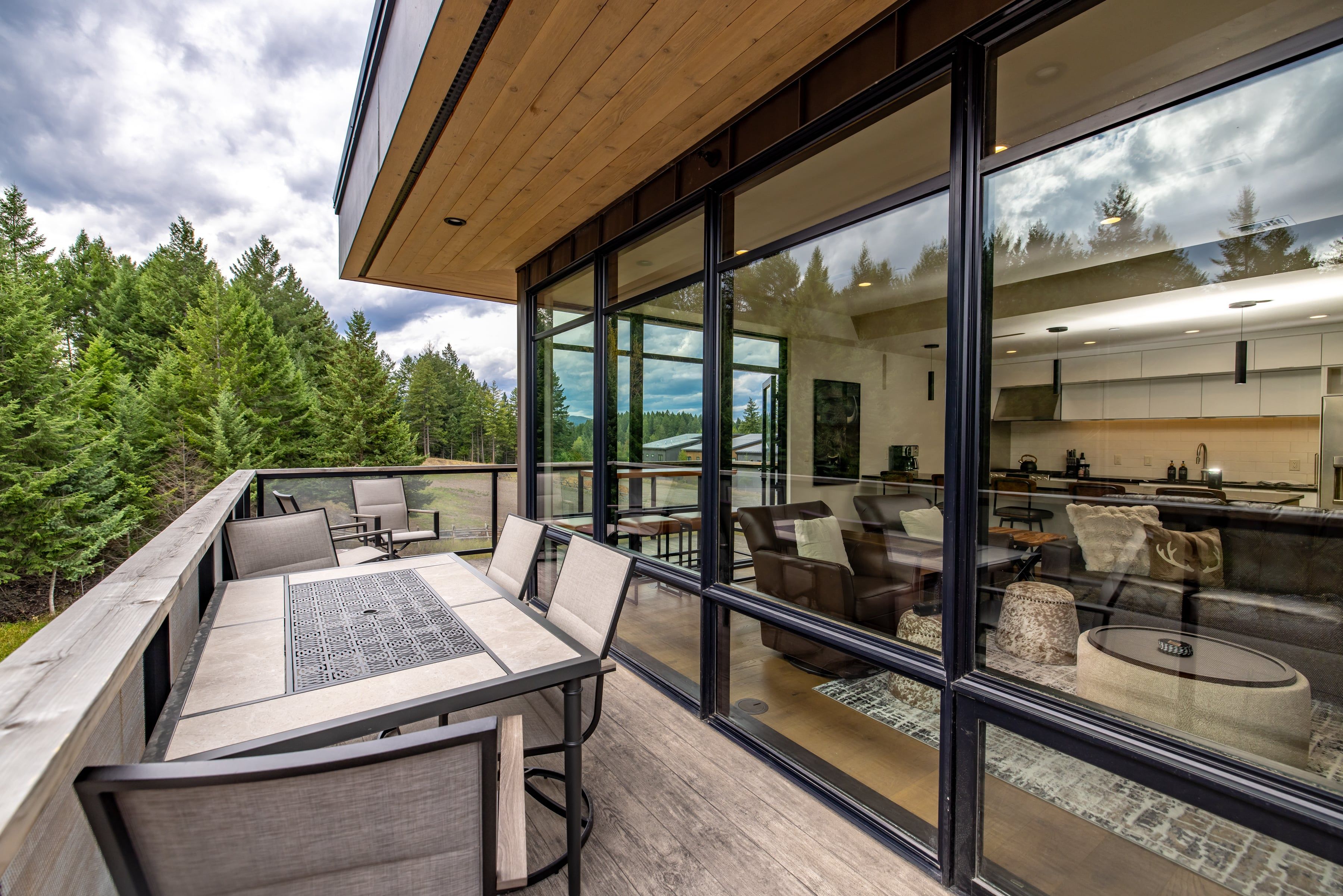 A modern outdoor dining area with a wooden deck, cushioned seating, and a glass wall offering a view of a forested landscape.