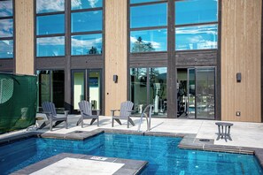 A swimming pool with lounge chairs and exercise equipment, surrounded by a building with large windows and wooden paneling. - Top-floor corner condo l Hot Tub / Summer Pool / Gym (Whitefish)