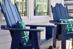 Zwei Adirondack-Stühle mit grün-weißen Karbokissen, auf einer Veranda mit Blick auf ein Gebäude durch ein Fenster platziert.