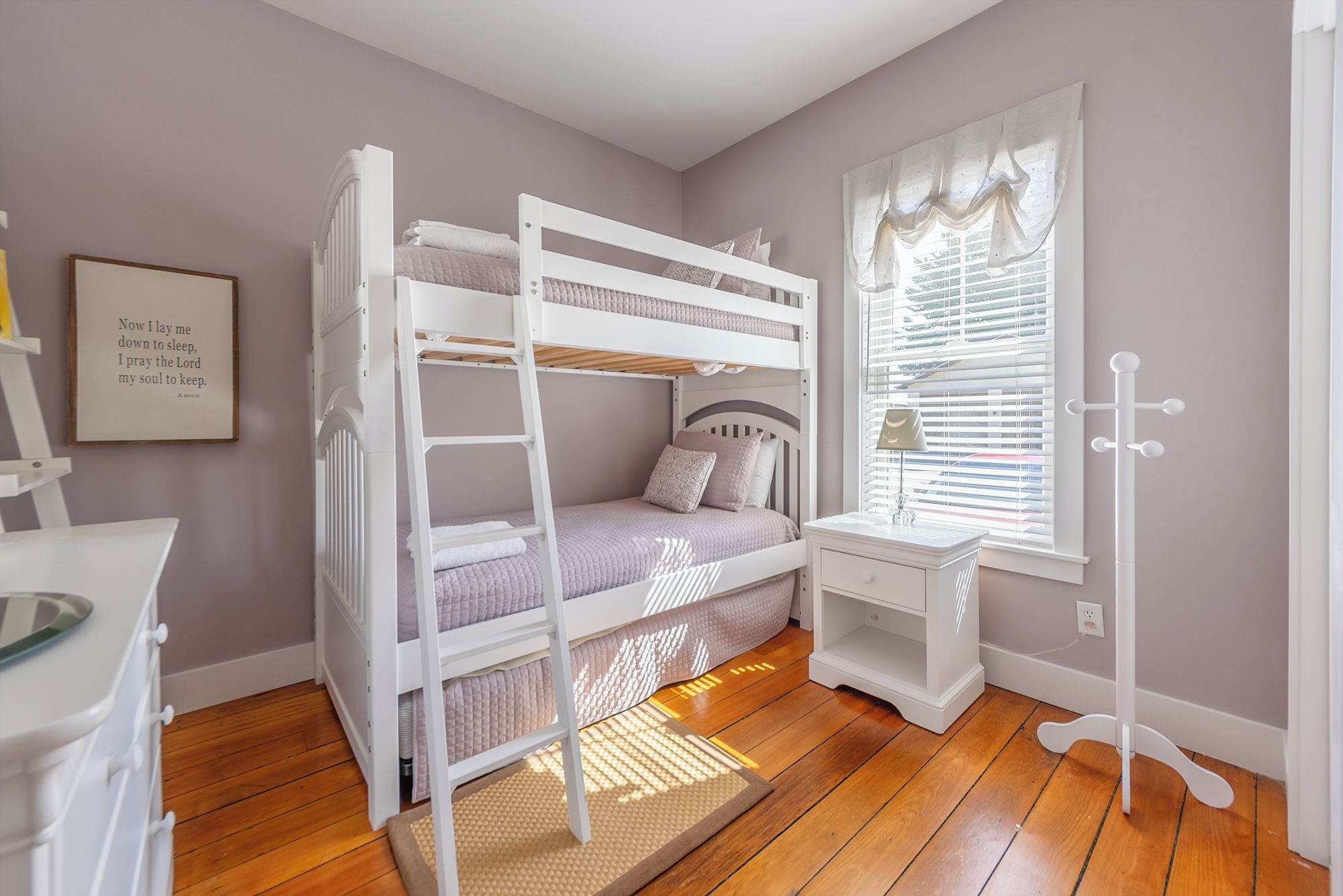 A child's bunk bed with a ladder, a desk with a chair, a window with blinds, and a coat rack.
