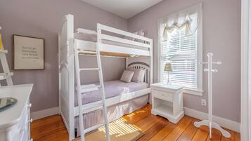 A child's bunk bed with a ladder, a desk with a chair, a window with blinds, and a coat rack.