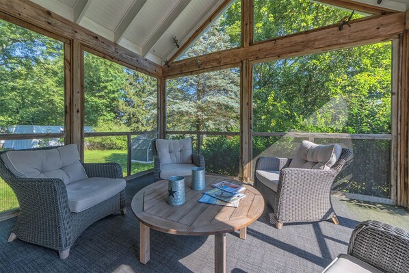 A covered outdoor seating area with wicker furniture and a round table, surrounded by a wooden railing.