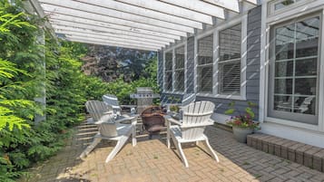 Eine Terrasse mit Gartenmöbeln und Grill, umgeben von viel Grün und einem Haus mit großen Fenstern.