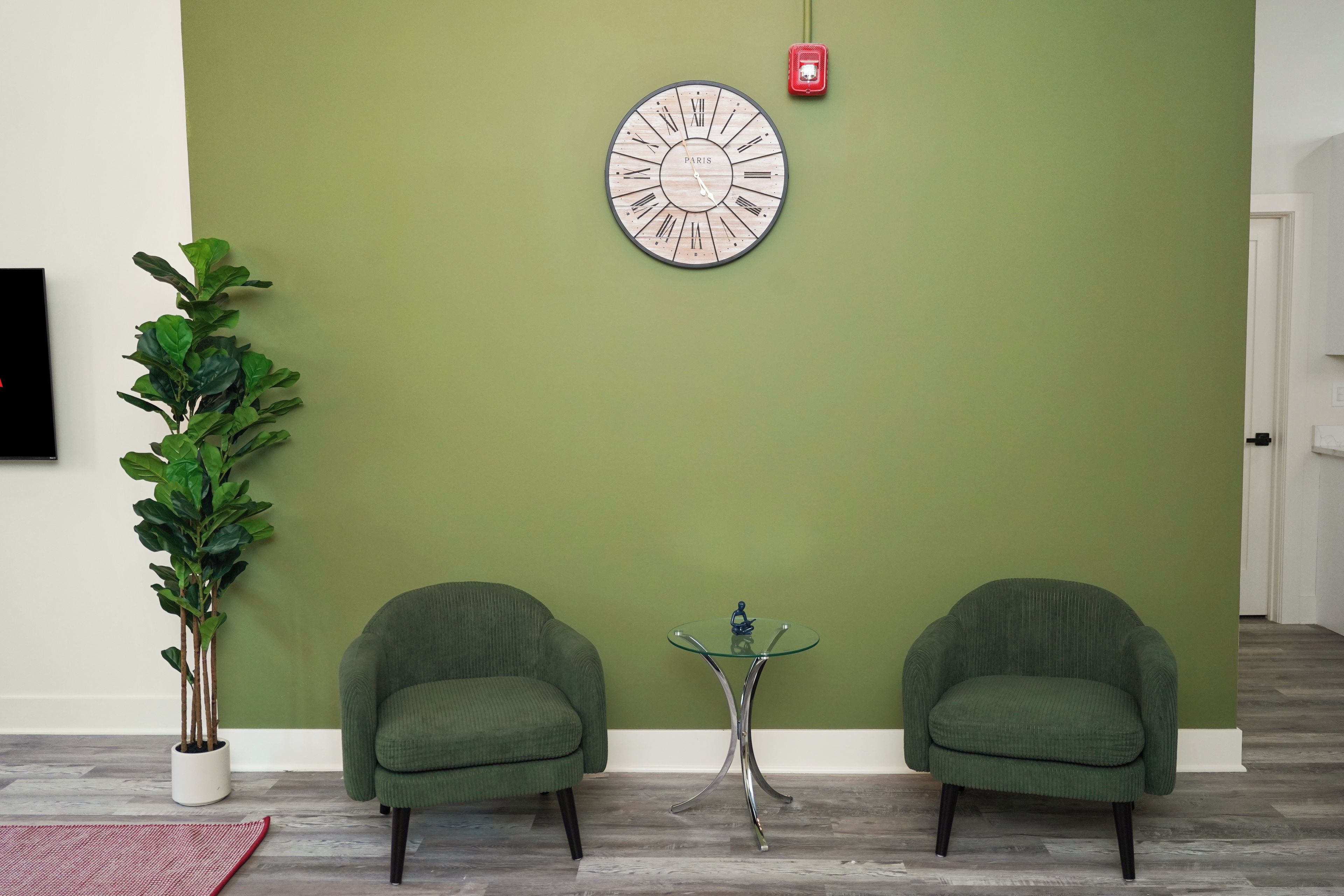 A room with a green wall, two green armchairs, a round wall clock, a potted plant, and a small table with a decorative item.