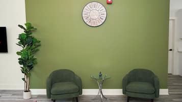 A room with a green wall, two green armchairs, a round wall clock, a potted plant, and a small table with a decorative item.