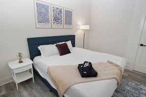 A neatly made bed with a white duvet, a dark blue headboard, and a single red pillow. A small bedside table with a globe and a white lamp is visible.