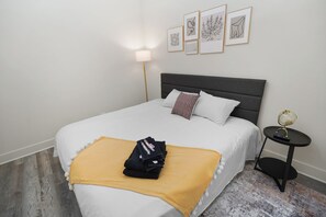 Una cama bien hecha con cubrecama blanco y manta amarilla, una mesita de noche con un globo terráqueo y control remoto, y una pared con cuadros enmarcados.