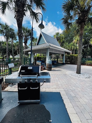 A grill station with a metal roof and a bench, surrounded by palm trees and a gazebo. - Cozy Modern Condo located in a beach Club in Kissimmee 15min from Disney (Kissimmee)