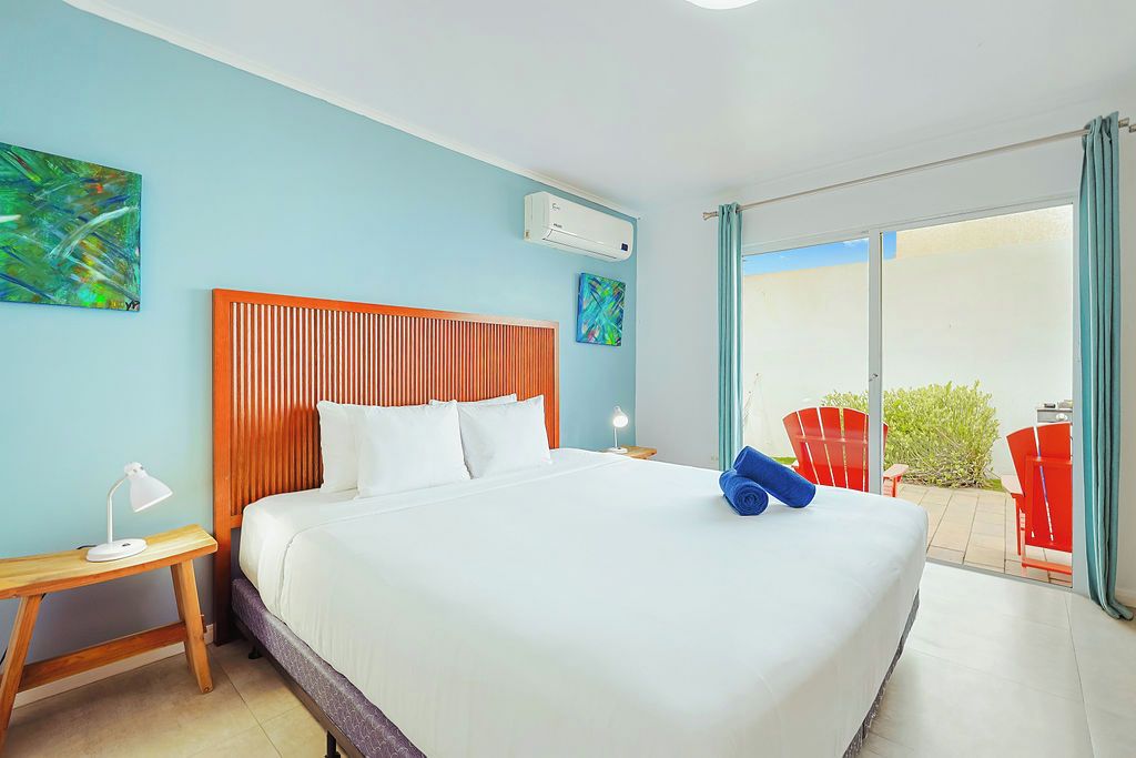 A hotel room with a large bed, a wooden headboard, a nightstand with a lamp, and a view of the outside patio with red chairs.